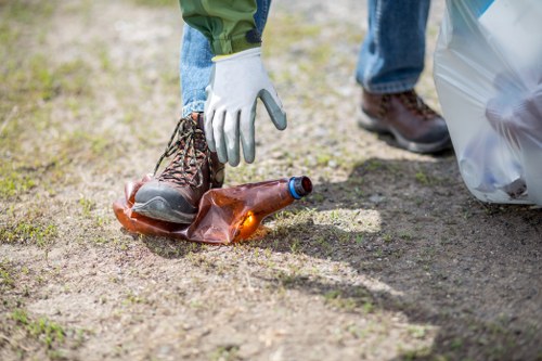 Close-up of a person wearing a white work glove, placing a used glass bottle into a clear plastic rubbish bag outdoors on a gravel surface. The individual is dressed in green trousers and sturdy brown work boots. In the background, another person in blue jeans and dark shoes is partially visible, standing nearby. The scene appears to be part of a rubbish or waste clearance activity, with the gravel area suggesting a driveway or garden setting. The focus is on the action of disposal, highlighting waste management practices that Leyton House Clearance might facilitate in local residential areas near Leyton, UK, as indicated by the postcode inferred from the page context. The lighting is natural, emphasizing the clarity and detail of the bottle, glove, and surrounding environment, underscoring the professional approach to rubbish collection and recycling efforts.