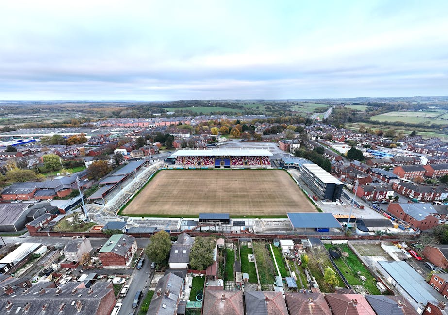 An aerial view of a football stadium situated within a residential and commercial area, with a large rectangular pitch in the center showing patches of brown grass and worn turf. Surrounding the pitch