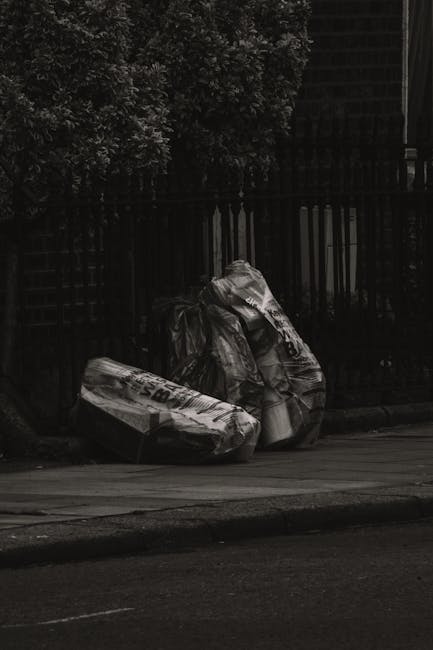 Three large black garbage bags filled with mixed waste sit on the pavement next to a black metal fence and a brick wall, partly shaded by nearby trees. The bags are crumpled and appear to contain hous