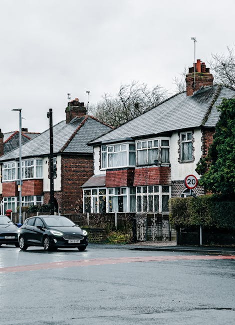 A residential street scene featuring two semi-detached houses with brick and white render facades, pitched tiled roofs, and prominent brick chimneys. The front gardens are bordered by low metal fences